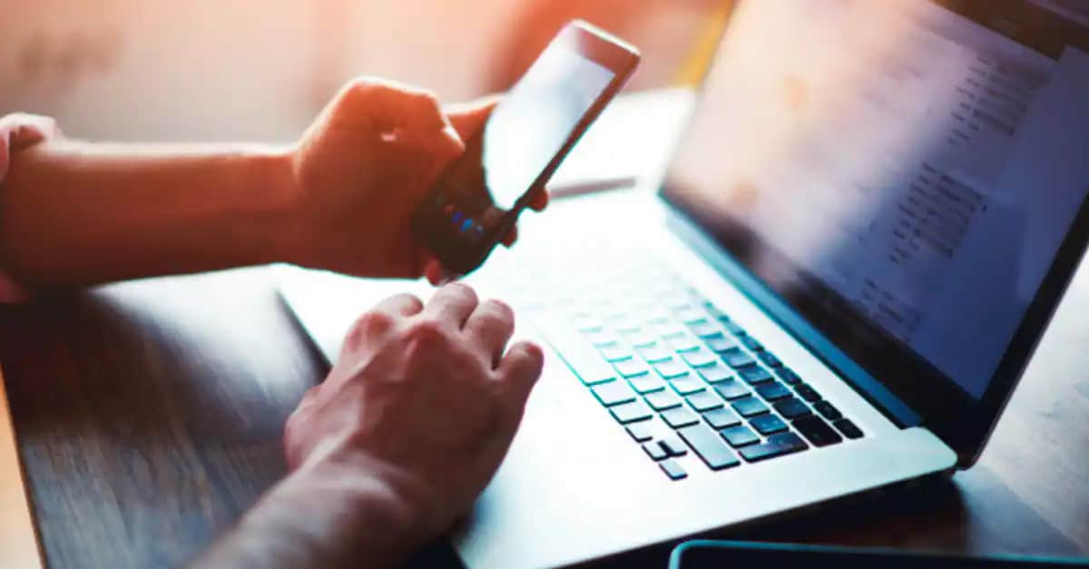 hand holding a mobile phone with a laptop on a table